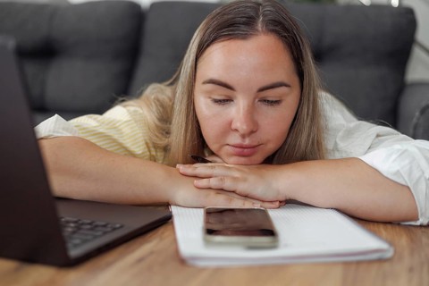 Young Caucasian woman with blond hair resting her head on folded arms at a table with a laptop and