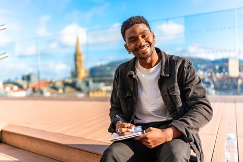 Happy african man writing notes sitting on a terrace with city views in a sunny day