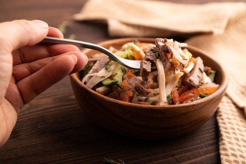 Meat salad with beef and vegetables in wooden bowl with hand on brown wooden background and linen