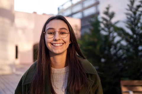 A woman with long brown hair and glasses smiles warmly outdoors She's wearing a green coat and