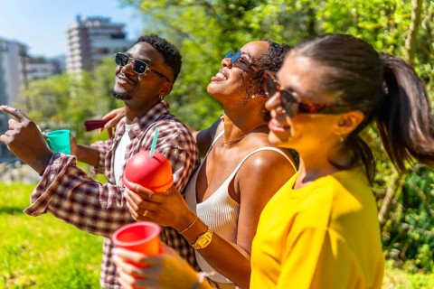 Three friends are having fun in a park, holding drinks and pointing at something in the distance