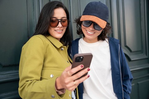A joyful lesbian couple shares a moment while looking at a phone outside. Their clothing and