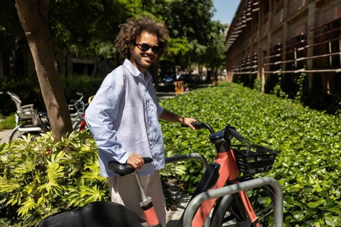 A cheerful Afro man in casual attire enjoys a sunny day outdoors, standing with a public bicycle,