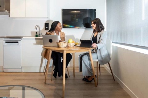 Two businesswomen are sitting at a table in a hotel room, working on their laptops and tablets