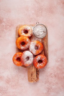 A group of baked donuts sits on a wooden cutting board. Powdered sugar is dusted on the donuts.