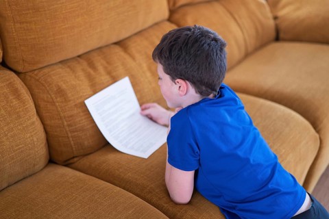 A boy in a blue shirt attentively reads a sheet of paper while sitting on a comfortable couch