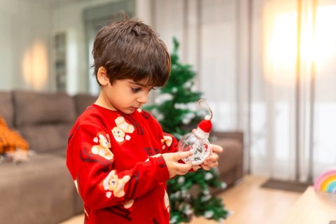 Young boy in cozy pajamas studies a clear christmas ornament topped with a tiny santa hat as he