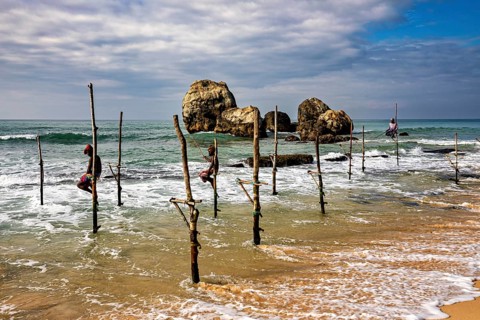 Several fishermen sit on wooden posts by the sea with large rocks in the background, The stilt