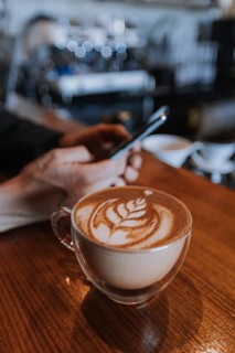 A man uses a smartphone at a cafe, focusing on a beautifully crafted latte with intricate art. The