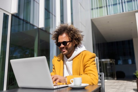 A businessman with afro hair, wearing sunglasses and a yellow coat, enjoys working on his laptop