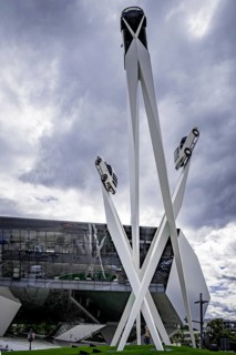 Modern sculpture of cars on high pillars in front of a modern building under a cloudy sky,