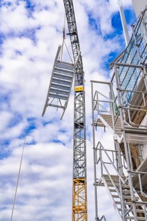 Crane lifts metal staircase into the air next to a scaffolding, in front of a cloudy sky, staircase