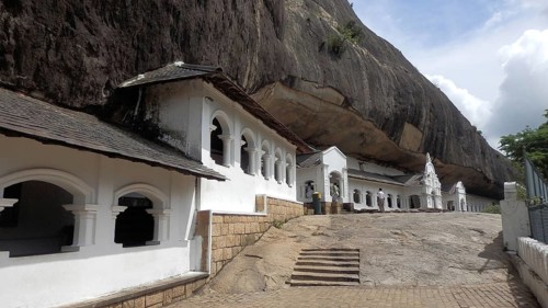 Rock temple with white architecture under a large rock wall in daylight, Dambulla cave temple, Sri