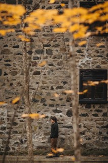 A person walks alongside a rustic stone building, surrounded by the warm hues of autumn leaves in