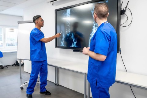 Two medical professionals wearing blue scrubs discussing a patient's spinal x rays displayed on a