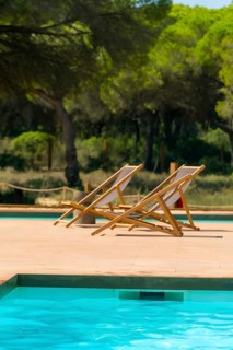 Two empty deckchairs are placed on a wooden deck next to a tranquil turquoise pool, surrounded by