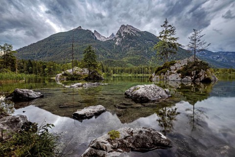 Mountain landscape with lake surrounded by trees and rocks, dramatic clouds over the Alps, The