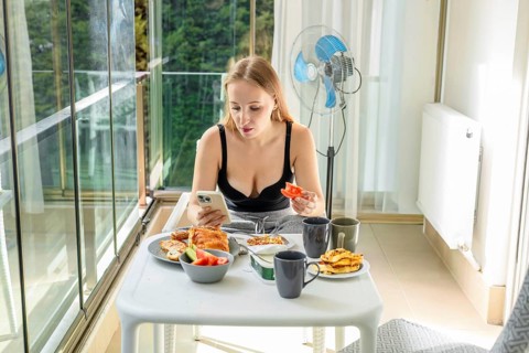 A young Caucasian woman with long brown hair sits at a table on a balcony. She is using a