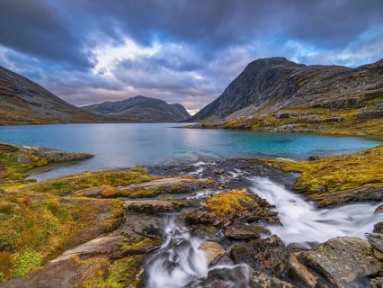 A mountain lake with a small waterfall surrounded by rocks and a dramatically cloudy sky, sunset