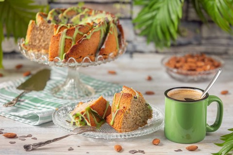 A sliced cake and pieces of cake with green icing next to a cup of coffee and almonds