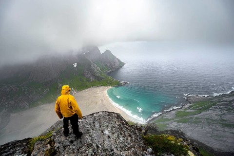 Hiker on Helvetestinden summit, view of Bunesstranden sandy beach and blue sea, dramatic cloudy