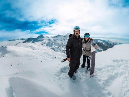 Smiling couple posing together with a snowboard on a snowy mountain summit, overlooking vast peaks