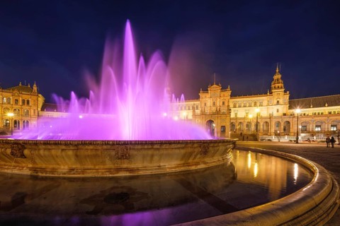 Purple illuminated fountain and curved basin stand before the glowing central pavilion of Plaza de
