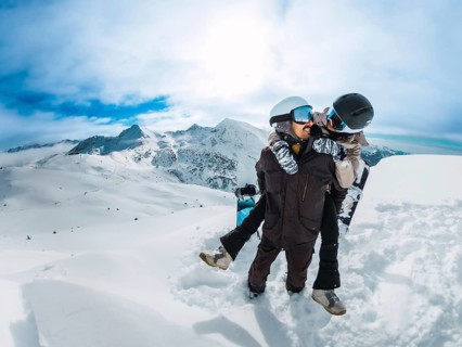 Snowboard couple sharing a playful piggyback on a snowy mountain peak, smiling and embracing winter