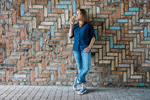 Woman drinking coffee while leaning against patterned brick wall, A woman in casual attire stands