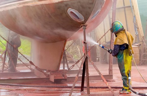 Worker sandblasting a sailing yacht hull in full protective clothing and helmet, creating a cloud