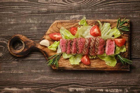Sliced beef steak placed on wooden cutting board with lettuce and tomato. This meal is presented on