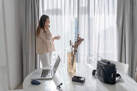 Woman standing by sheer curtains talking on smartphone and holding water glass, laptop and camera