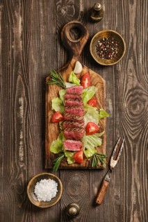 Beef steak cut into pieces placed on wooden cutting board. Plate includes lettuce, tomatoes, garlic