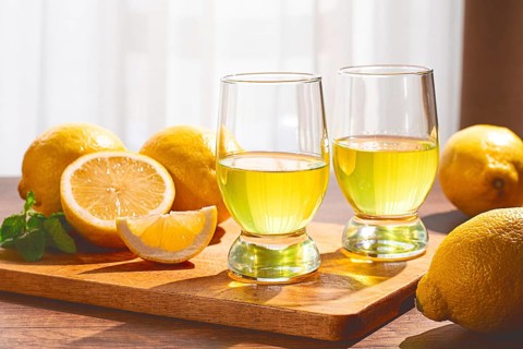 Two clear glasses sit on a wooden board filled with yellow lemon drink. Whole and sliced lemons are