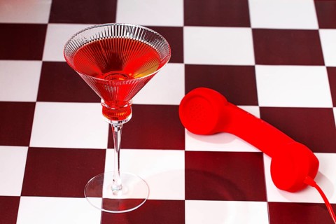 A red telephone rests beside a cocktail glass filled with red liquid. This scene takes place on a