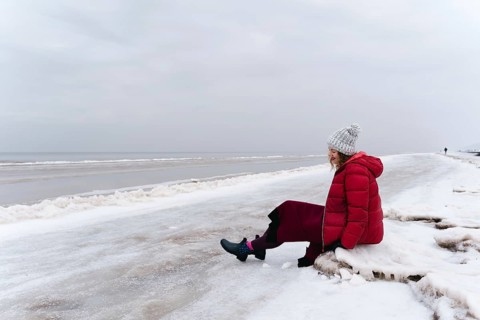 A cheerful woman in a red jacket and a warm dress sits on the shore of the winter sea, A woman in a