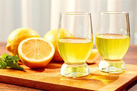 Two clear glasses hold yellow liquid on a table. Fresh lemons are placed beside the glasses. One