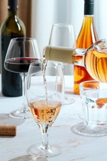 A person is pouring rose wine into a clear glass. There are several wine bottles and other glasses