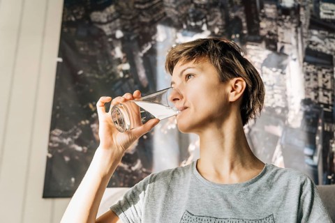 A young woman with short hair drinks water from a glass, Woman in a gray shirt drinks water from a