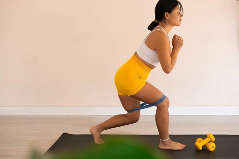 Woman lunging with a resistance band on a mat next to dumbbells indoors