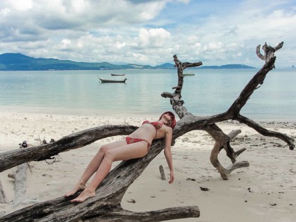 A woman in a red bikini reclines on a large weathered driftwood tree on a sandy tropical beach.