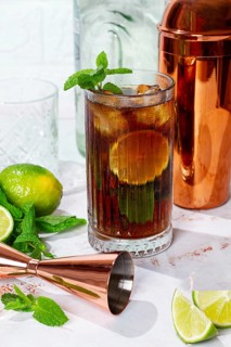 A tall glass filled with a dark soda drink shows slices of lime and fresh mint on a white table. A