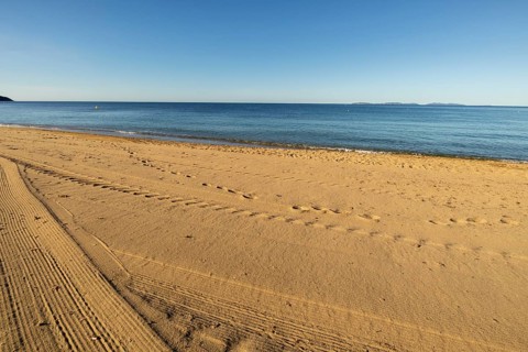 Beach-level viewpoint across raked sand with footprints, calm sea, distant islands, and clear