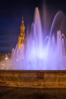 White and lavender fountain jets frame the illuminated North Tower of Plaza de Espana in a vertical