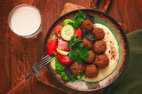 A plate holds falafel balls, tahini, salad with tomatoes and cucumbers, and a drink. The setting is