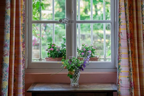 Flower vase standing on a wooden table in front of a window with a view of blooming plants outside,