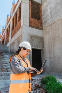 Woman in a hard hat and safety vest uses a laptop on a construction site. The background features