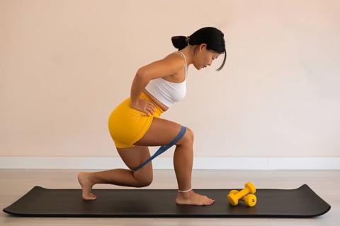 Woman lunging with resistance band next to dumbbells on an exercise mat indoors