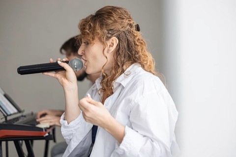 A young Caucasian woman with curly hair sings into a microphone at a home concert. A man is seated