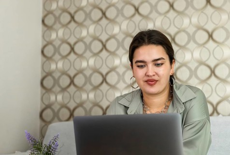 A young Hispanic woman with long brown hair works on a laptop at a wooden table in a cozy living